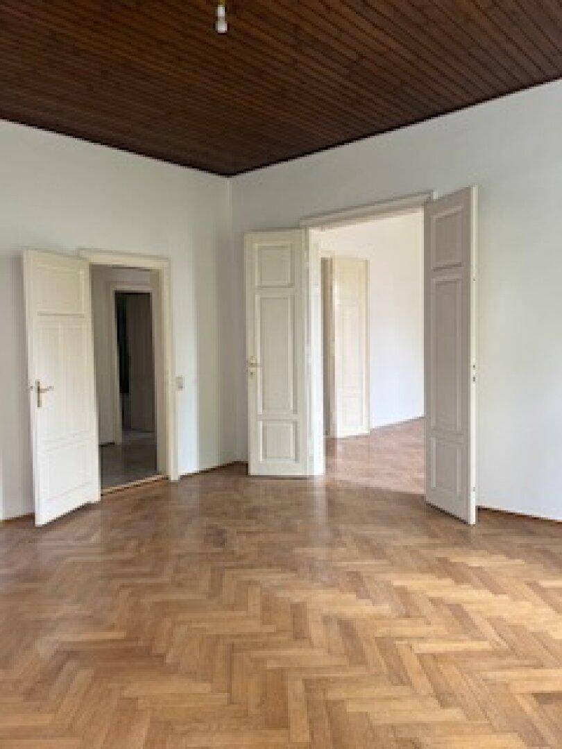Großes Wohnzimmer mit Fischgrätparkett und mehreren Türen zu angrenzenden Räumen.