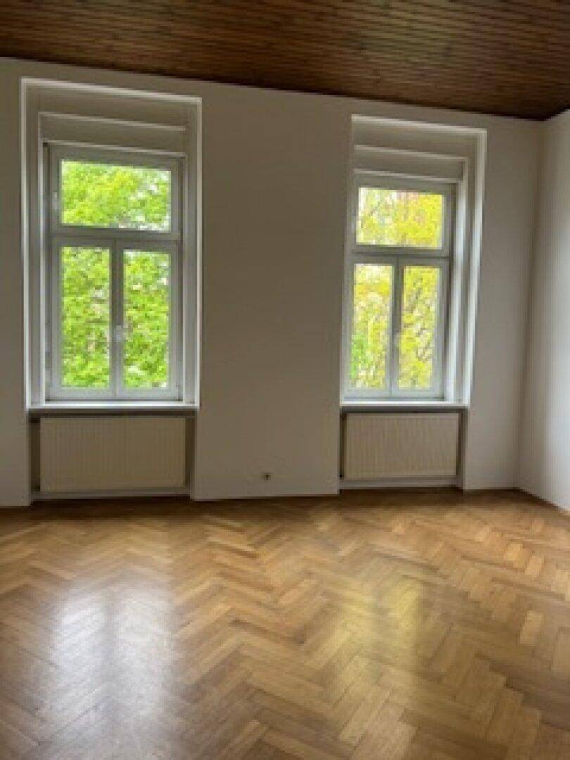 Helles Wohnzimmer mit zwei großen Fenstern, Fischgrätparkett und Holzbalkendecke.