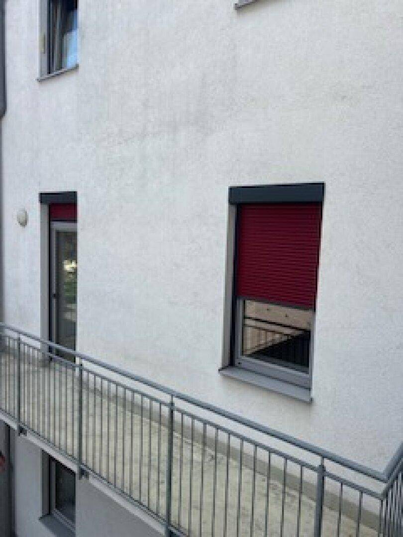 Weiße Gebäudefassade mit Fenstern, Rollläden und einem Balkon mit Metallgeländer.