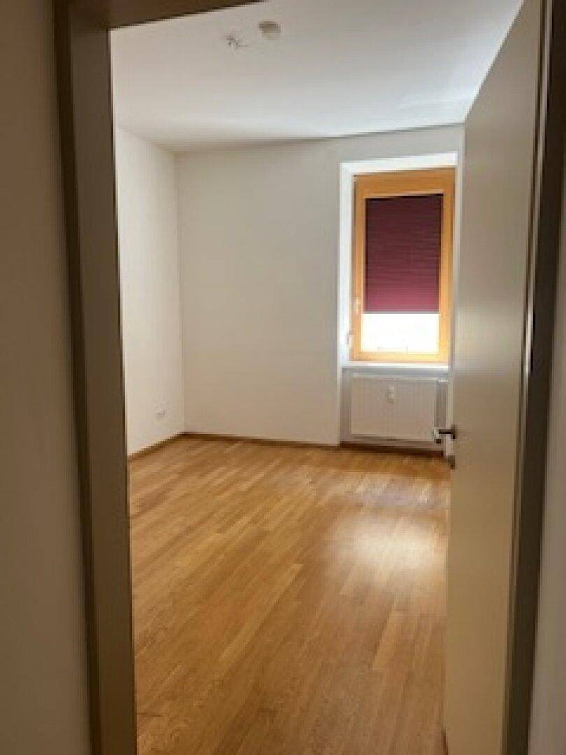 Leeres Zimmer mit hellem Holzboden, weißen Wänden und einem Fenster mit Rollladen.