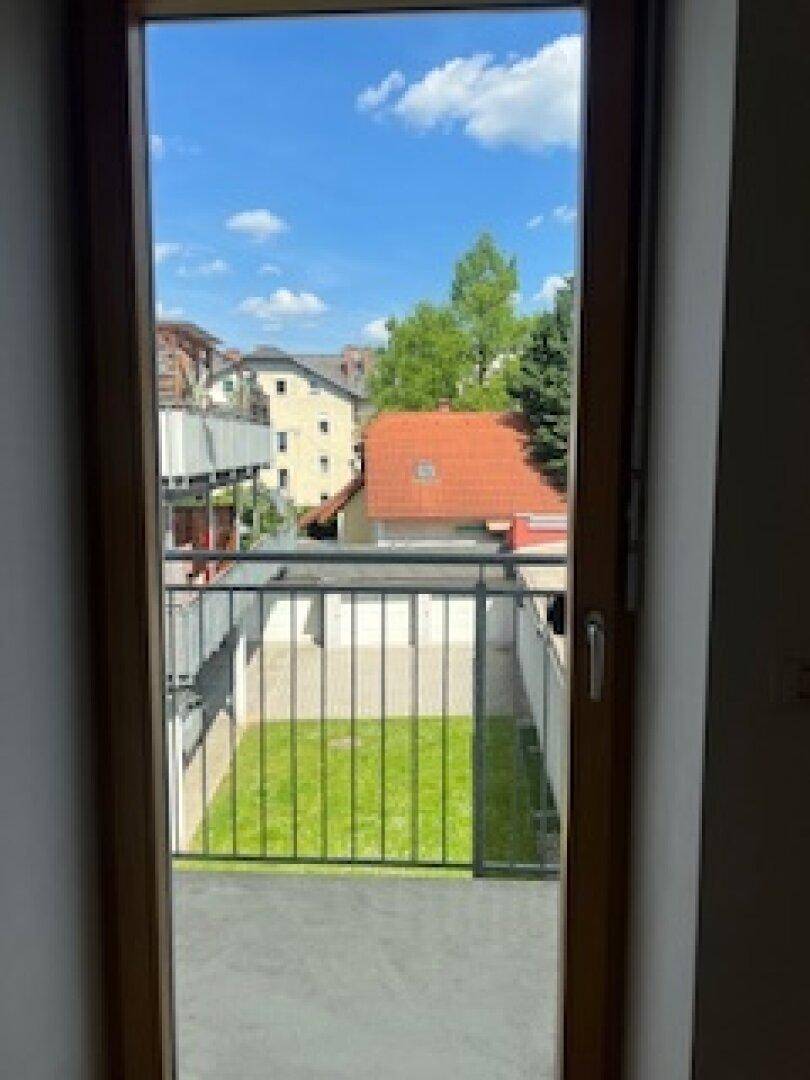 Blick vom Balkon auf einen grünen Innenhof und umliegende Gebäude unter blauem Himmel.