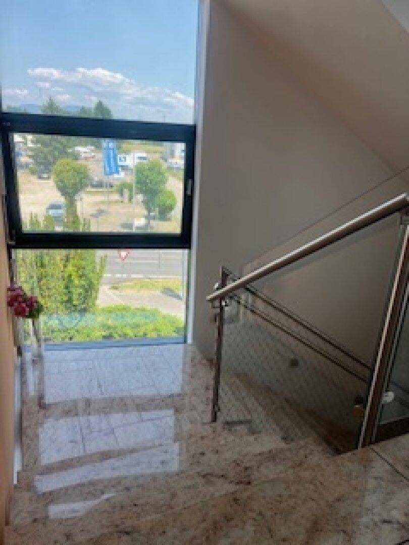 Treppenhaus mit Glasgeländer und großen Fenstern, die einen Blick auf die Straße und Bäume bieten.
