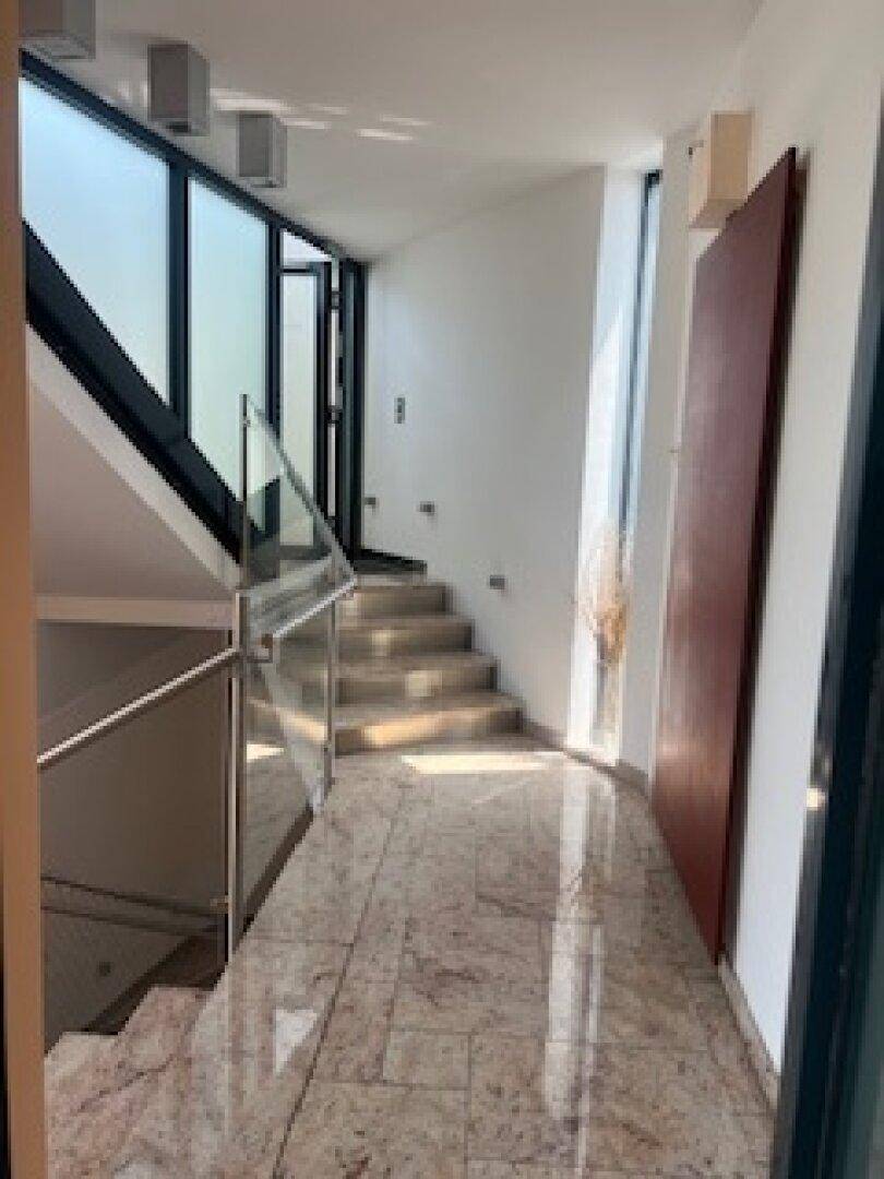 Heller Flur mit Treppe, Glasgeländer und großen Fenstern, die viel Tageslicht hereinlassen.