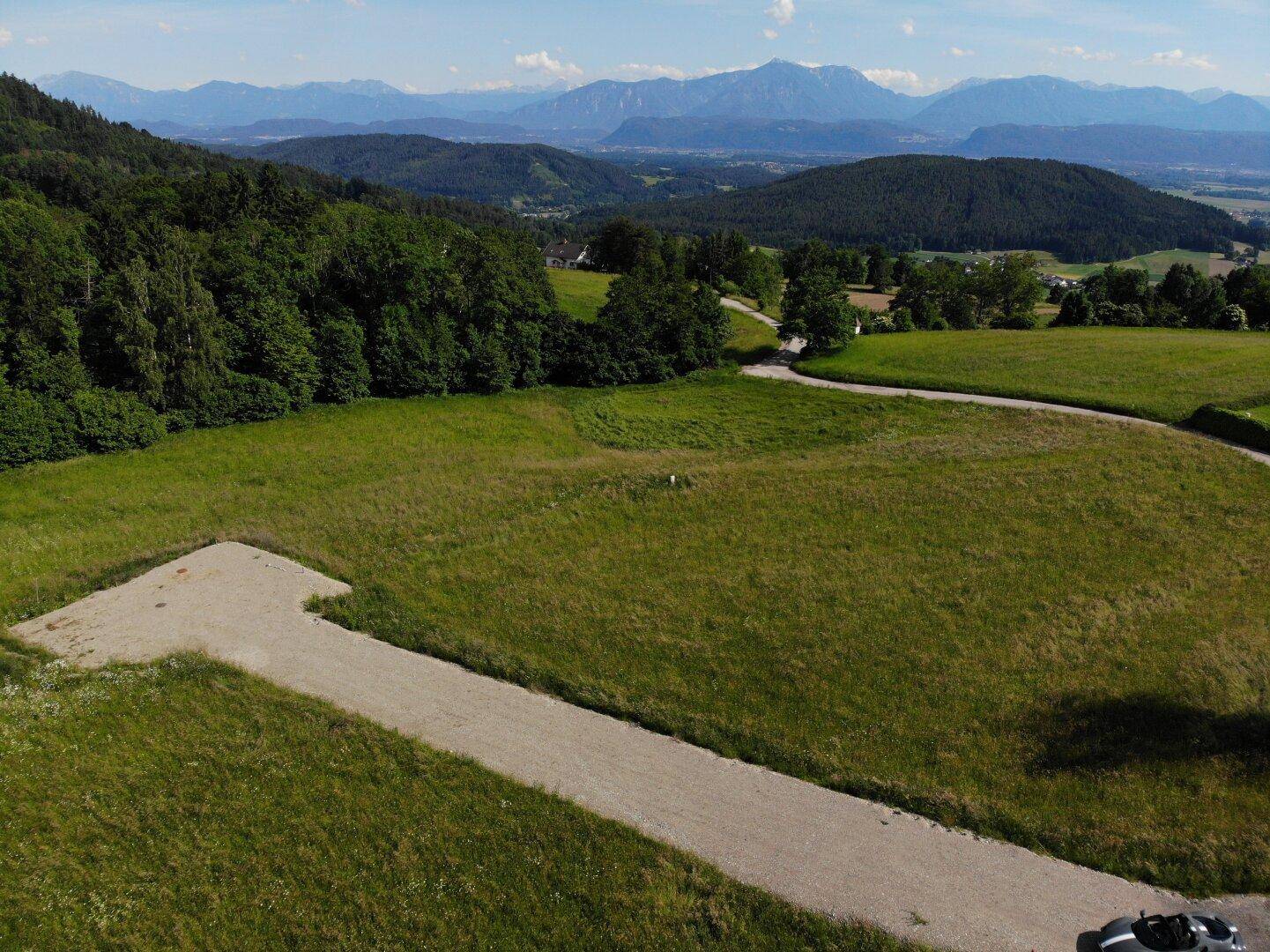 Weitläufiges Grundstück mit Zufahrtsweg, umgeben von grünen Hügeln und majestätischen Bergen am Horizont.