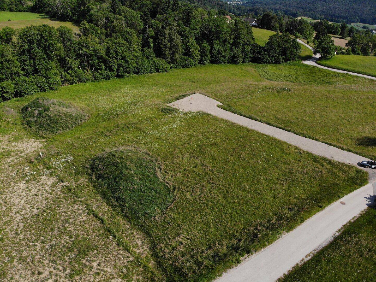 Luftaufnahme eines unbebauten Grundstücks mit Zufahrtsweg und angrenzendem Wald in ländlicher Umgebung.