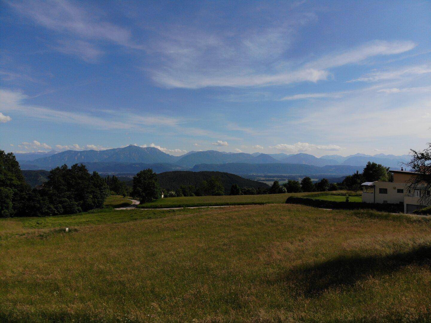 Weitläufige Landschaft mit grünen Feldern, Bergen im Hintergrund und einem modernen Haus.