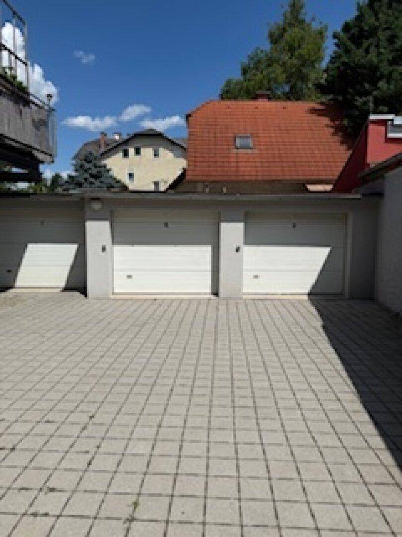 Mehrere Garagen mit weißen Toren und gepflastertem Vorplatz, dahinter ein Wohngebäude mit rotem Dach.