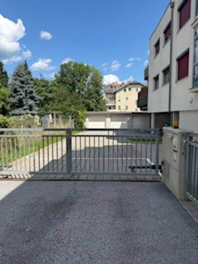 Eingangsbereich zu einer Garagenanlage mit Schiebetor und umliegenden Wohngebäuden unter blauem Himmel.