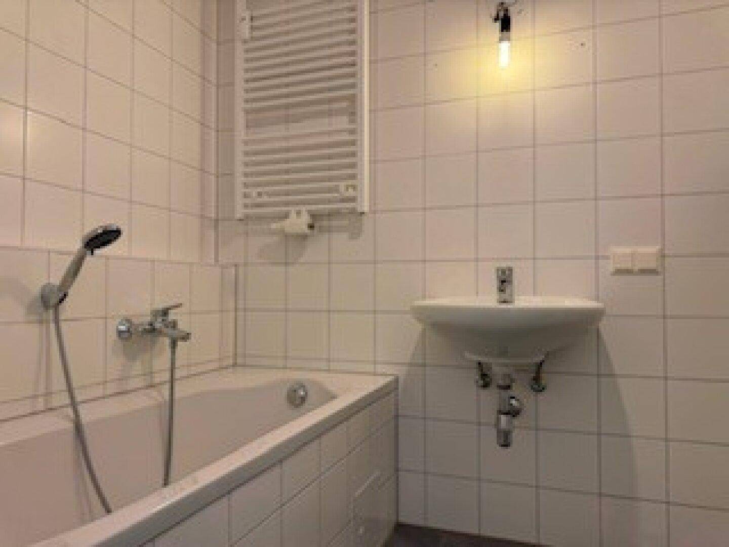 Helles Badezimmer mit weißen Fliesen, Badewanne, Waschbecken und einem beheizten Handtuchhalter.
