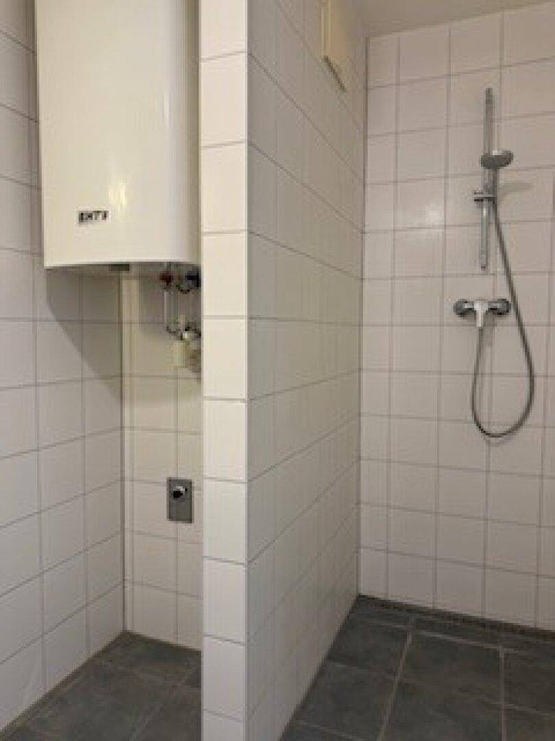Badezimmer mit weißen Fliesen, Duschbereich und einem Warmwasserboiler, ausgestattet mit dunklem Fliesenboden.