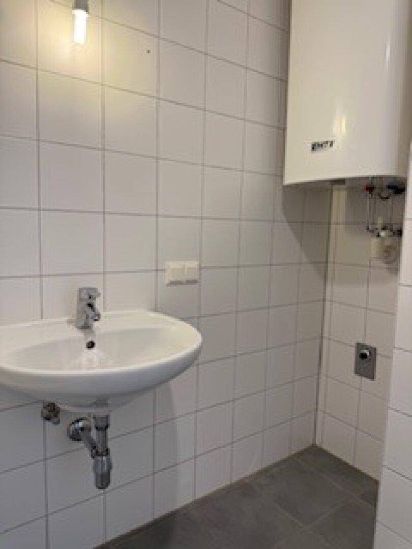 Badezimmer mit weißen Fliesen, Waschbecken und einem Warmwasserboiler, auf dunklem Fliesenboden.