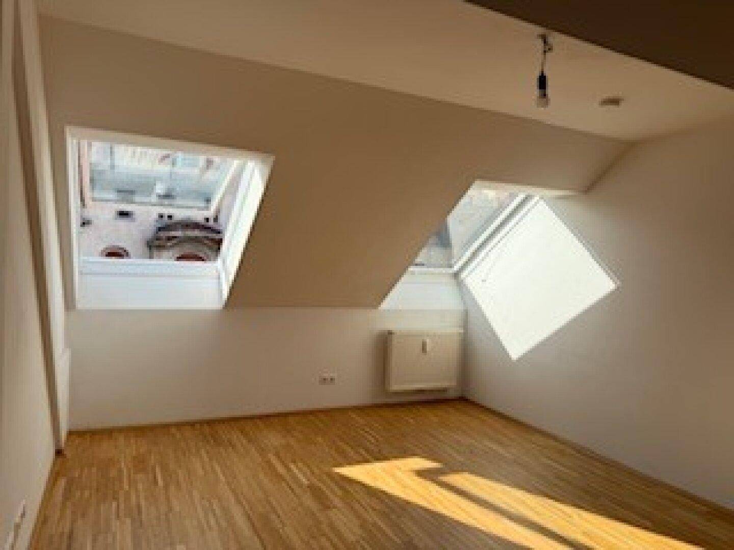 Helles Zimmer mit Holzboden, Dachschrägen und zwei Dachfenstern, die viel Sonnenlicht hereinlassen.