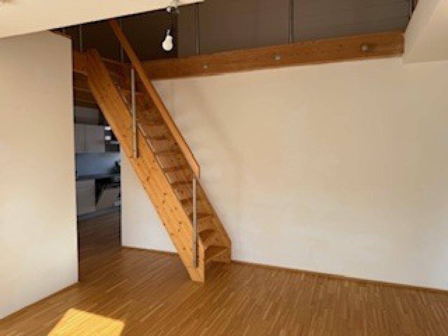Heller Wohnraum mit Holzboden, Dachschrägen und einer Holztreppe mit Metallgeländer.