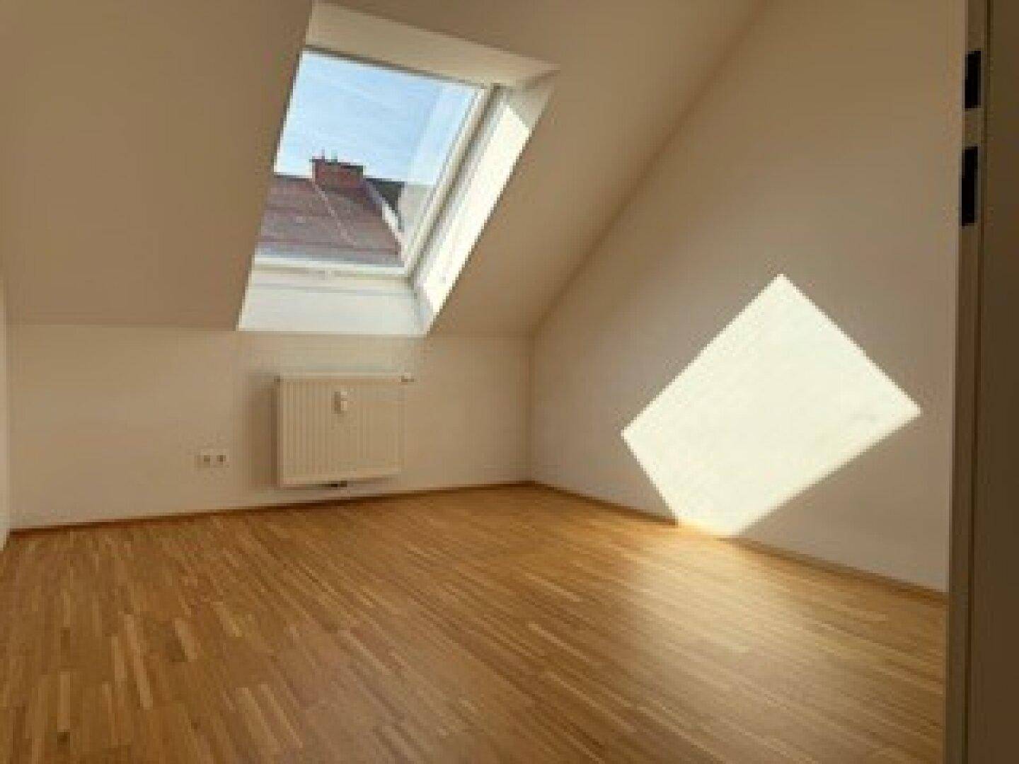 Helles Zimmer mit Holzboden, Dachschräge und einem großen Dachfenster, das viel Licht spendet.