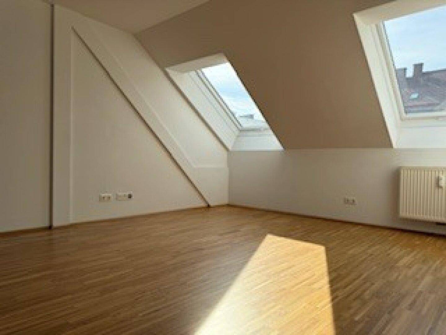 Geräumiges Zimmer mit Holzboden, Dachschrägen und zwei Dachfenstern, die für Helligkeit sorgen.