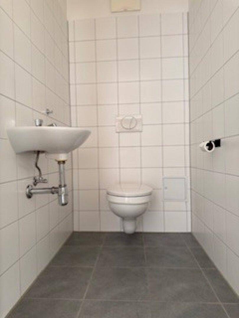 Modernes WC mit weißen Fliesen, wandhängender Toilette und kleinem Waschbecken, auf dunklem Fliesenboden.