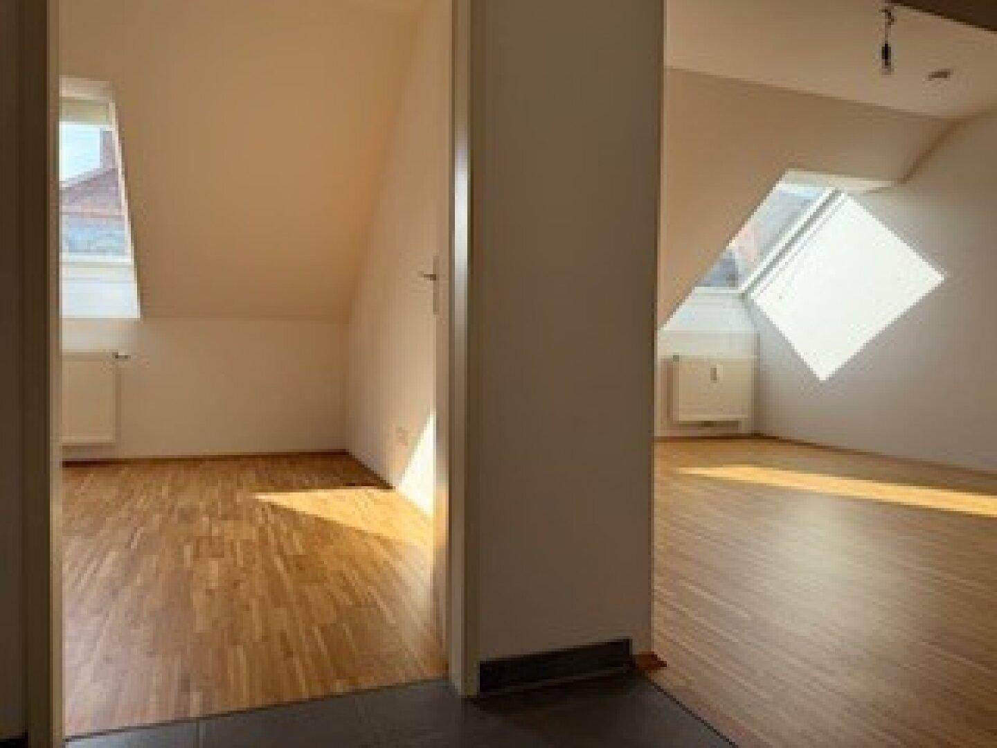 Flur mit dunklen Fliesen, der den Blick auf zwei helle Zimmer mit Holzböden und Dachfenstern freigibt.