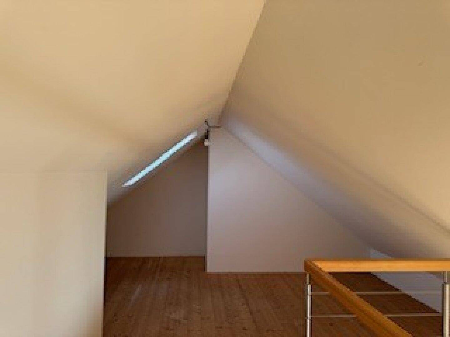 Heller Dachbodenraum mit schrägen Wänden, Holzdielenboden und einem schmalen Dachfenster.