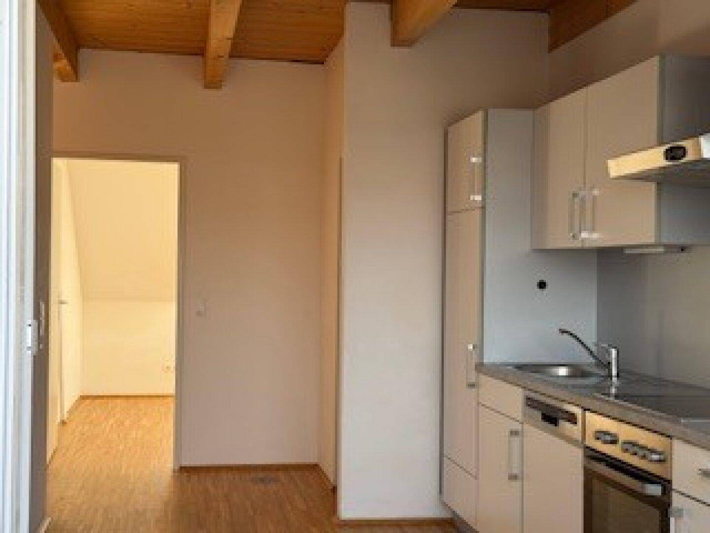 Helle Küchenzeile mit Holzboden und offenem Durchgang zu einem weiteren Zimmer, unter sichtbaren Holzbalken.