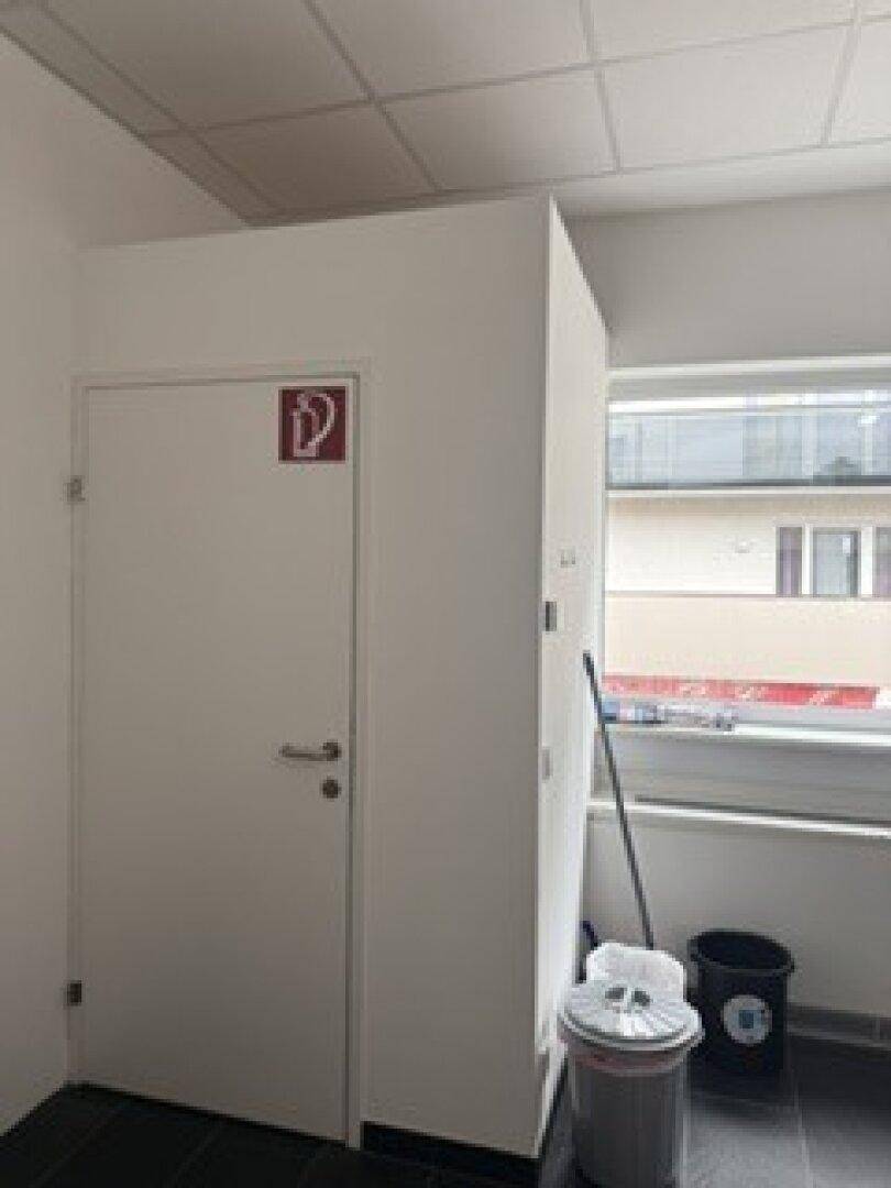 Abstellraum mit dunklem Fliesenboden, Fenster und einer weißen Tür mit Brandschutzzeichen für Sicherheit.