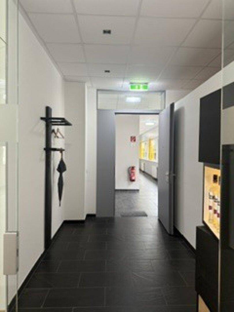 Langer Flur mit dunklem Fliesenboden, Garderobe und Blick auf einen Feuerlöscher für Notfälle.