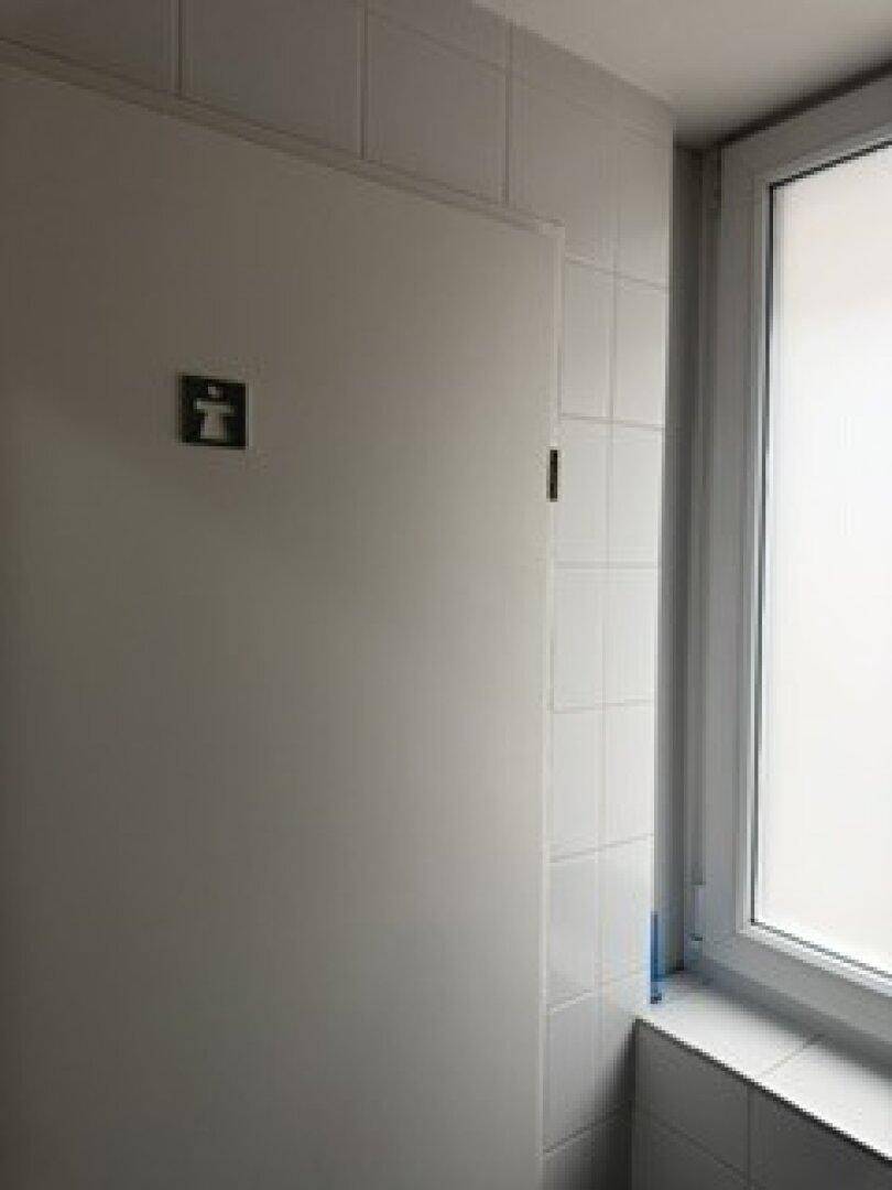 Toilettenkabine mit weißer Tür, Fliesen und einem Fenster, das für Belüftung sorgt.