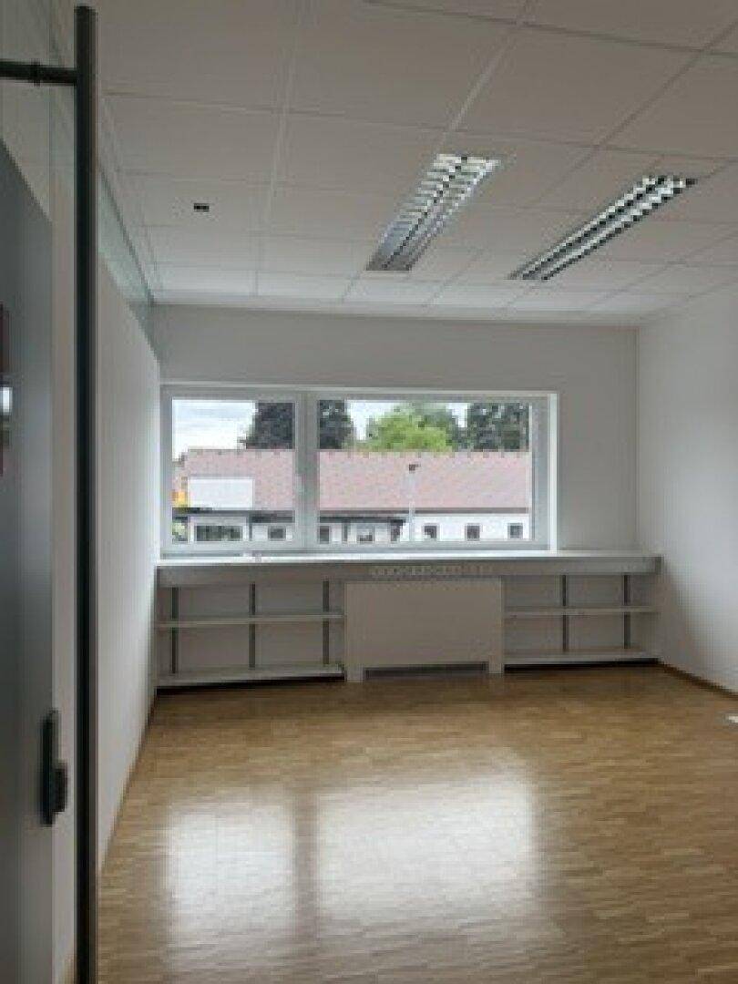 Leeres Büro mit Holzparkettboden, großen Fenstern und eingebauten Regalen für Stauraum.