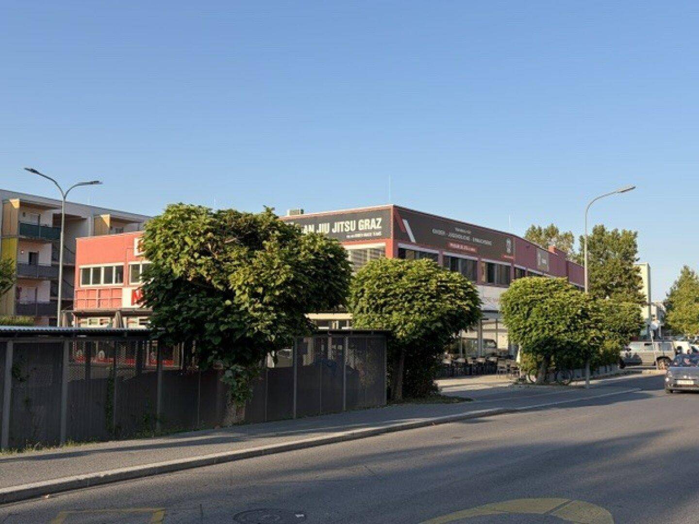 Seitenansicht des Geschäftsgebäudes von der Straße aus, mit Bäumen und einem angrenzenden Wohngebäude.