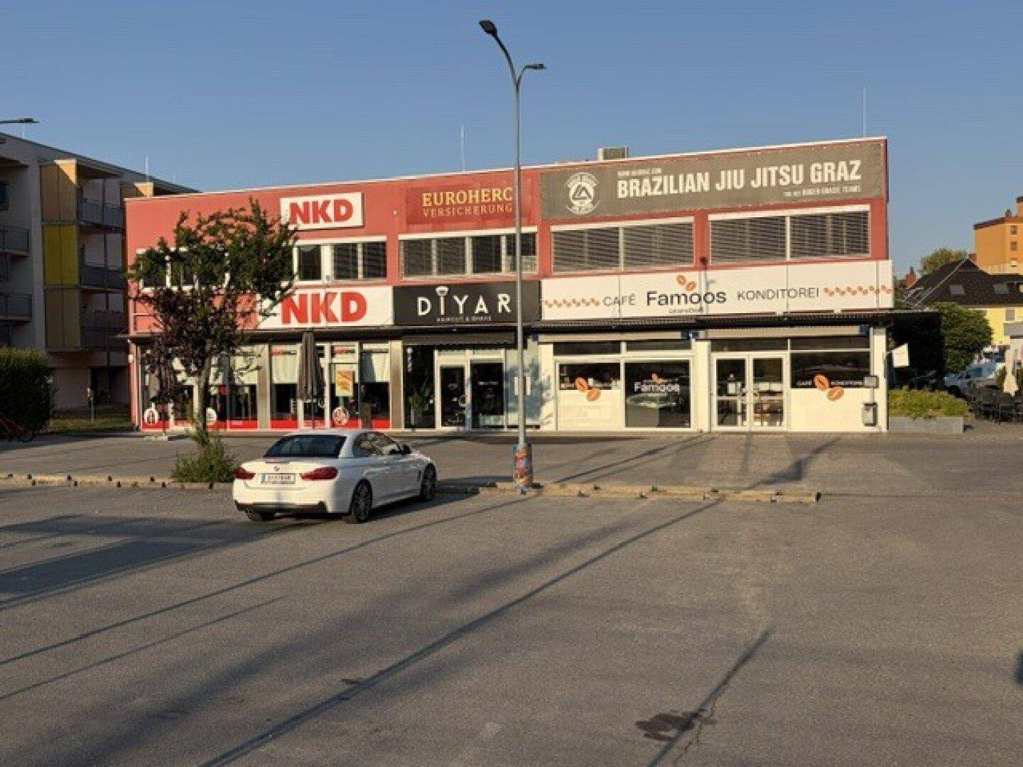 Breitere Ansicht des Geschäftsgebäudes mit Parkplatz im Vordergrund und verschiedenen Ladenfronten.