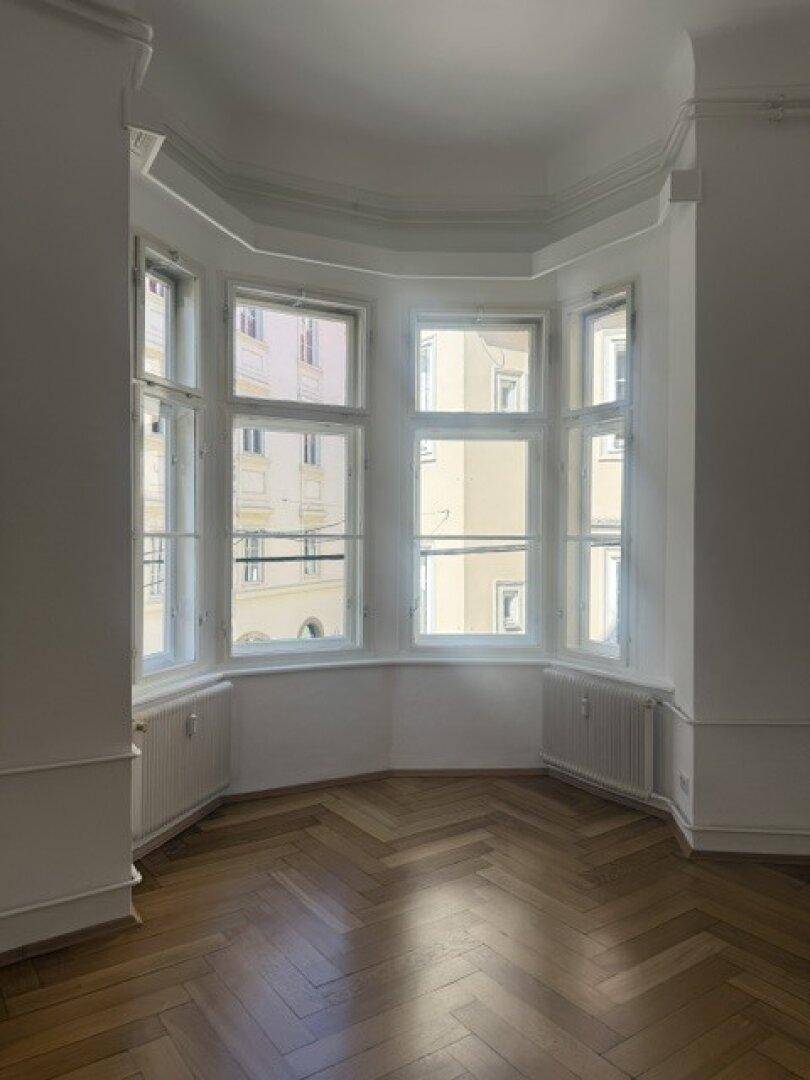 Heller Raum mit Fischgrätparkett und einem Erkerfenster, das eine charmante und helle Wohnatmosphäre schafft.