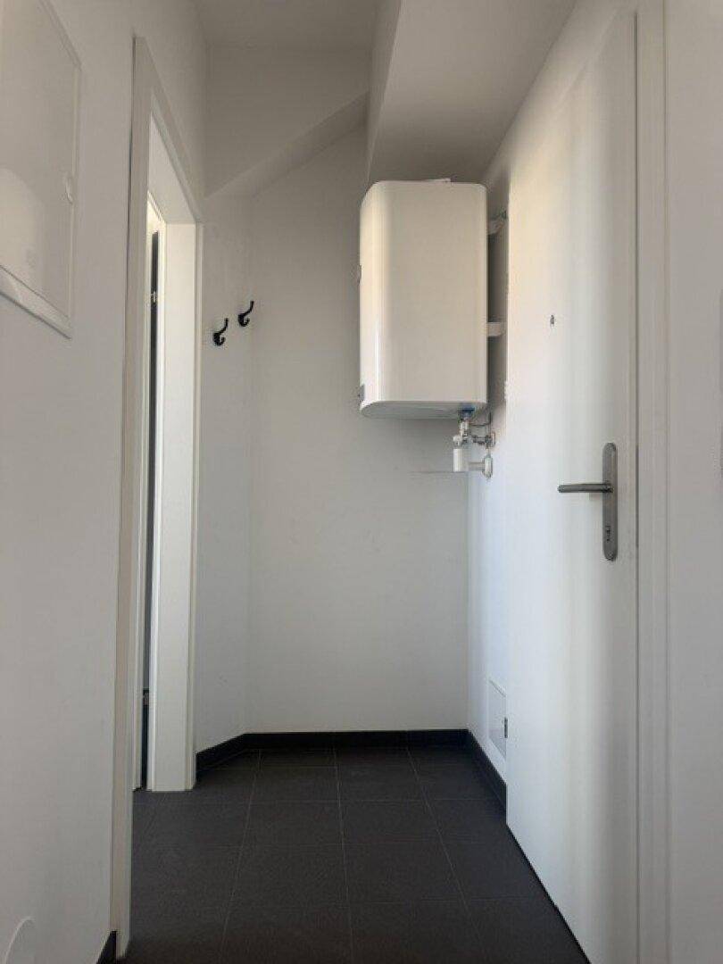 Schmaler Flur mit dunklen Fliesen, weißem Warmwasserboiler und Garderobenhaken an der Wand.