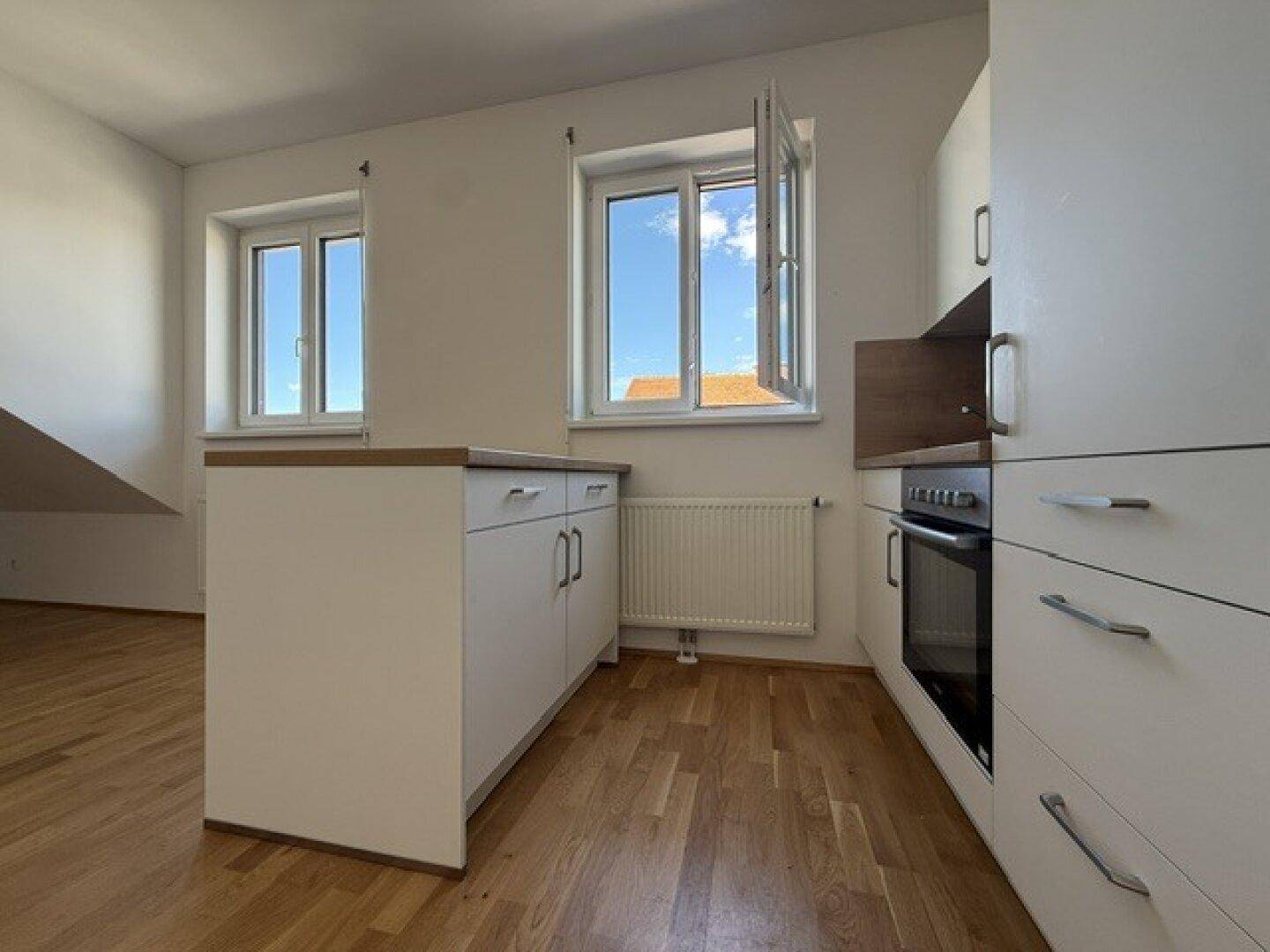 Offener Küchenbereich mit weißer Küchenzeile, Holzboden und zwei Fenstern mit Blick nach draußen.