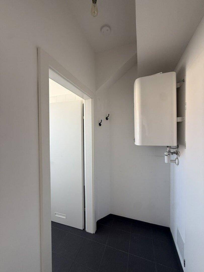 Flur mit dunklen Fliesen und einem Warmwasserboiler an der Wand, der zu einem weiteren Raum führt.