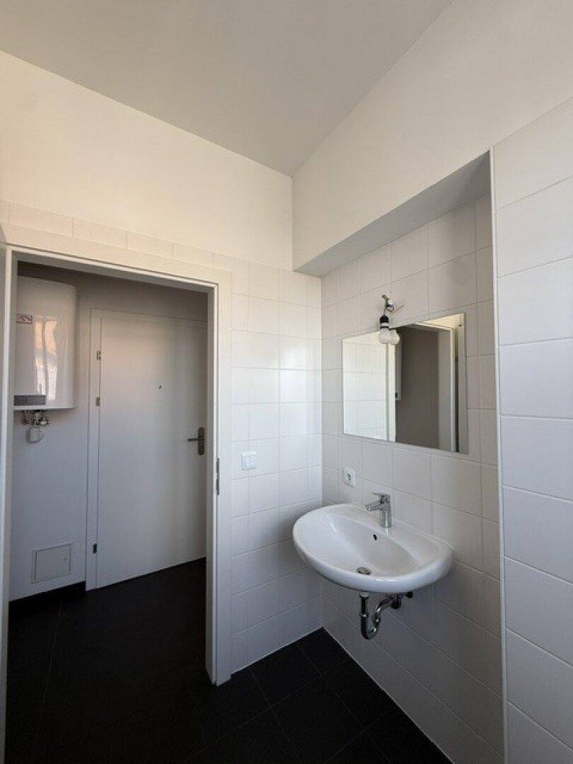 Badezimmer mit Waschbecken, Spiegel und Blick auf den Flur mit der Eingangstür.
