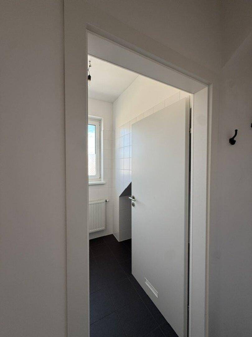 Badezimmerzugang mit offener Tür, die den Blick auf weiße Fliesen und ein Fenster freigibt.