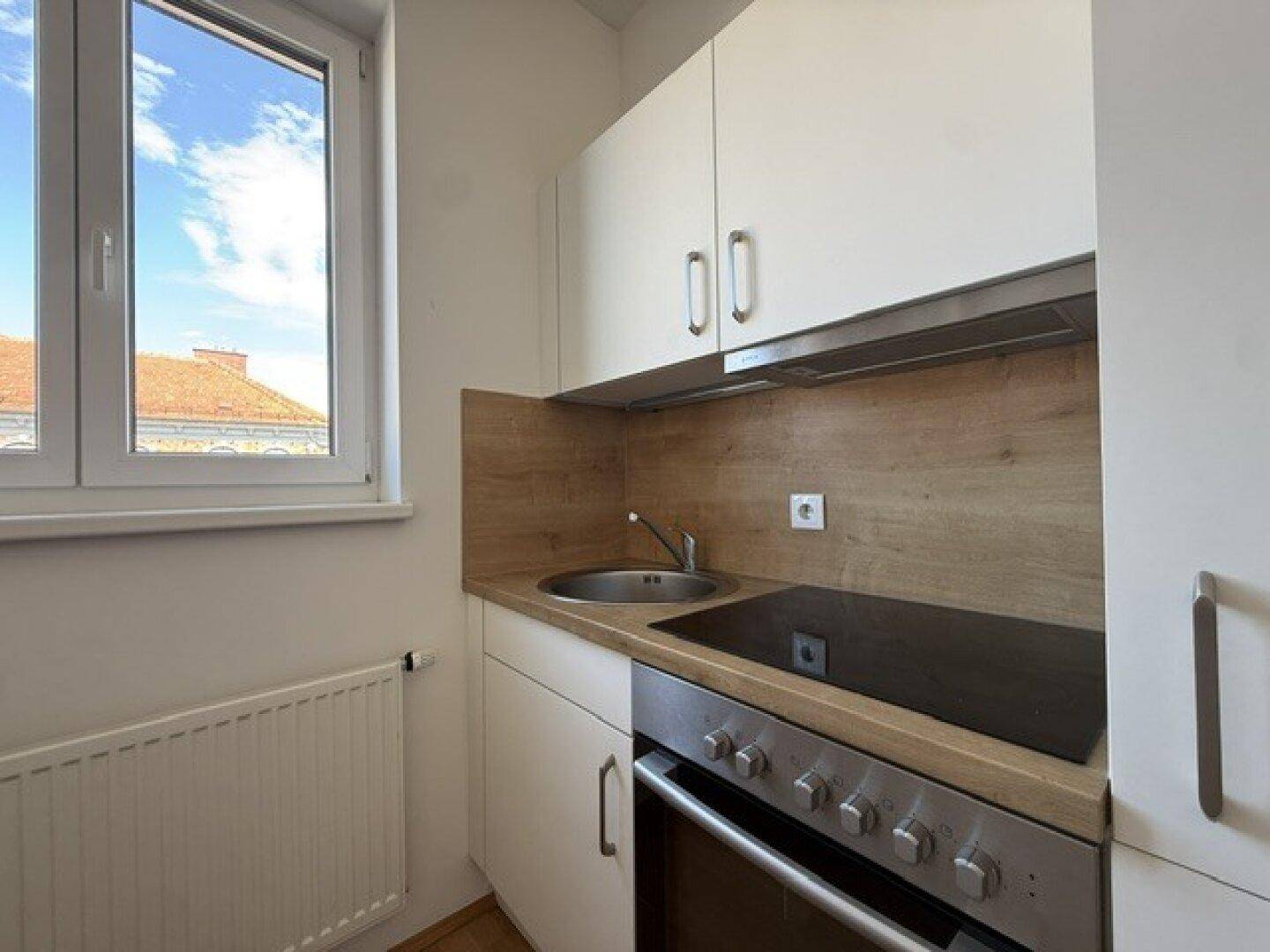 Kompakte Küchenzeile mit Kochfeld, Spüle und Fenster, das viel Tageslicht hereinlässt.