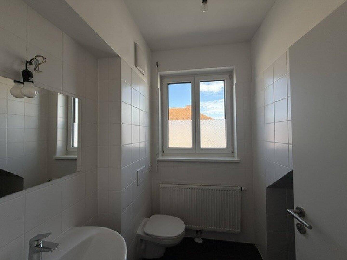 Helles Badezimmer mit WC, Waschbecken, Heizkörper und Fenster für natürliche Belüftung.