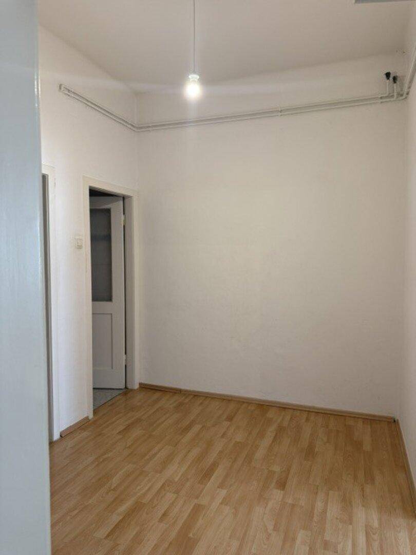 Leerer Raum mit hellem Holzfußboden, weißen Wänden und sichtbaren Heizungsrohren.
