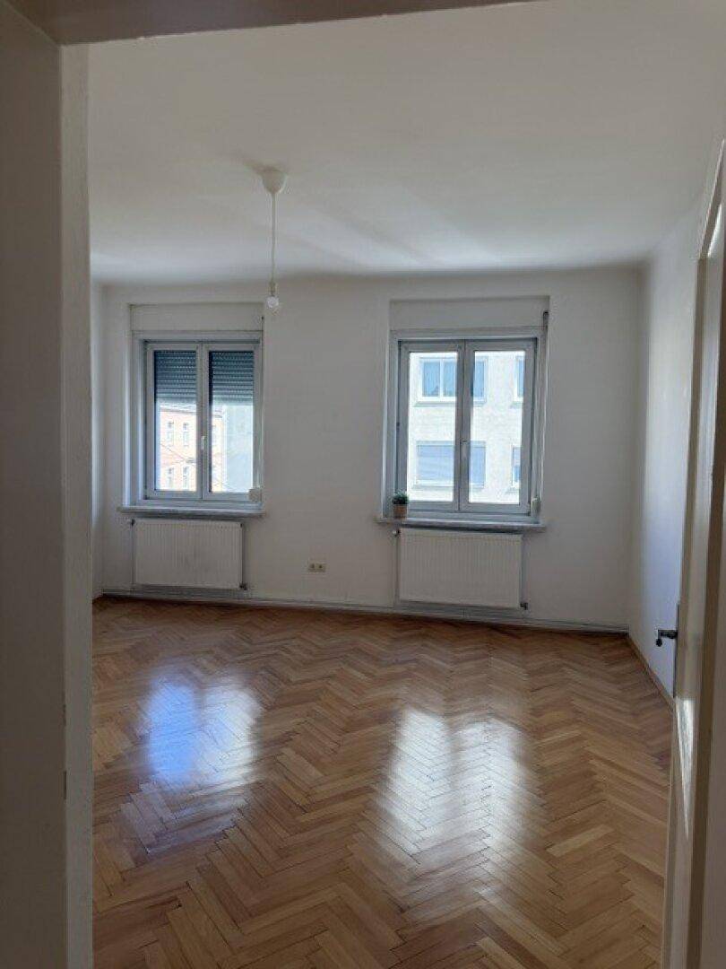 Geräumiger, heller Raum mit Fischgrätparkett und zwei Fenstern mit Heizkörpern.