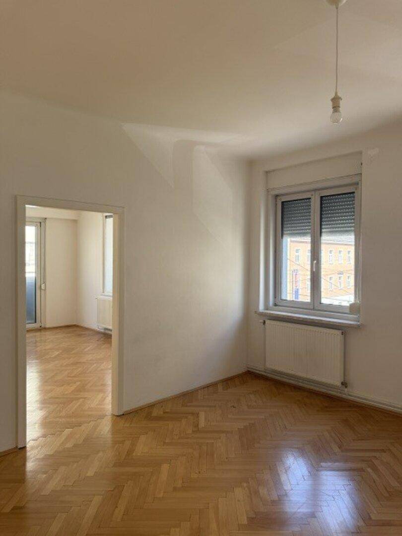 Heller Raum mit Fischgrätparkett, einem Fenster und Zugang zu einem weiteren Zimmer.