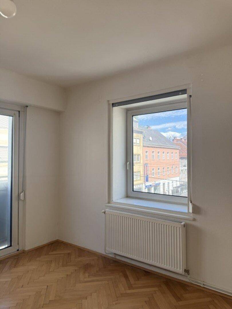 Helles Zimmer mit Fischgrätparkett, Heizkörper und Blick auf die Stadt durch das Fenster.