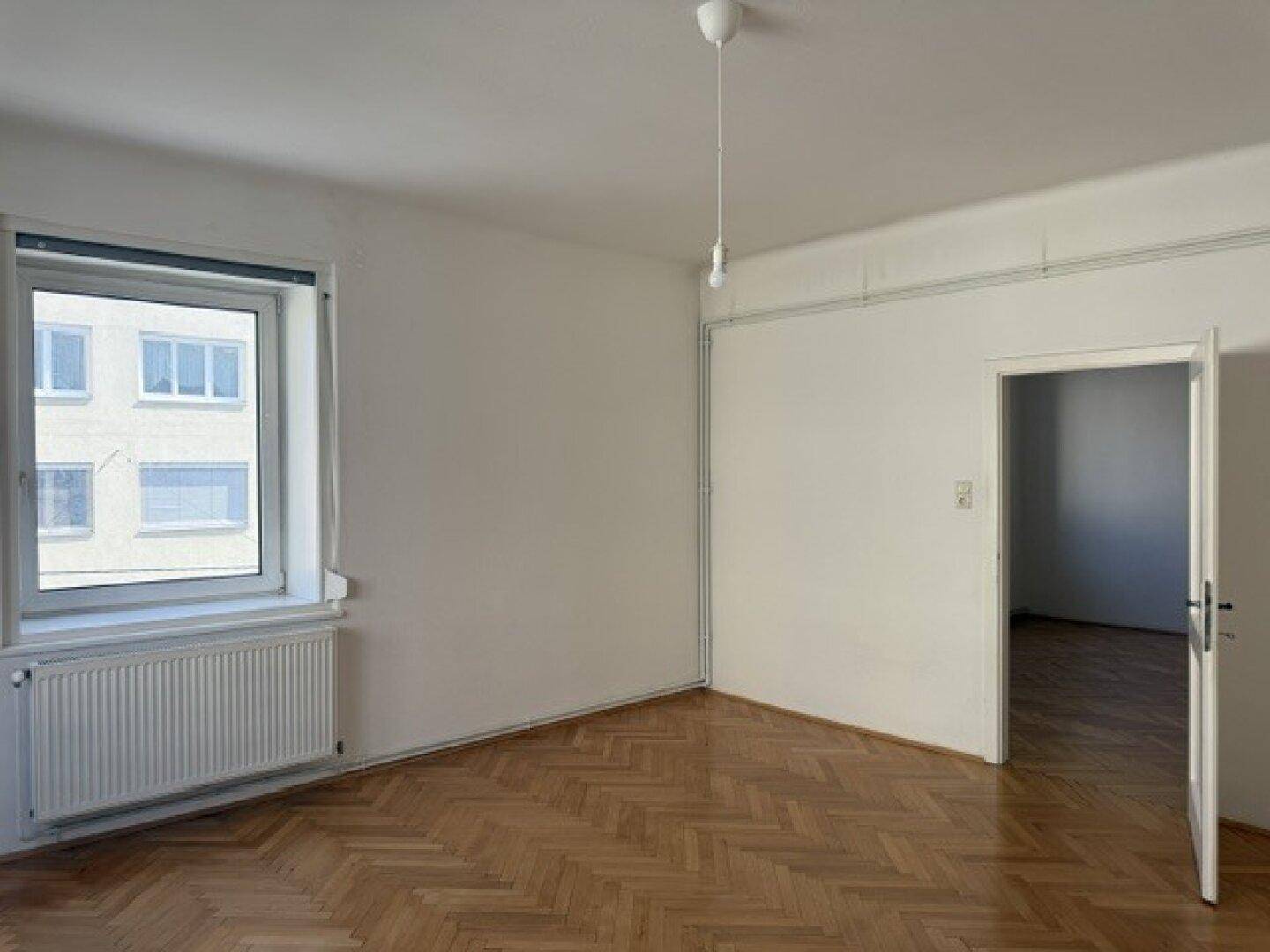 Geräumiges Zimmer mit Fischgrätparkett, Fenster und Zugang zu einem weiteren Raum.
