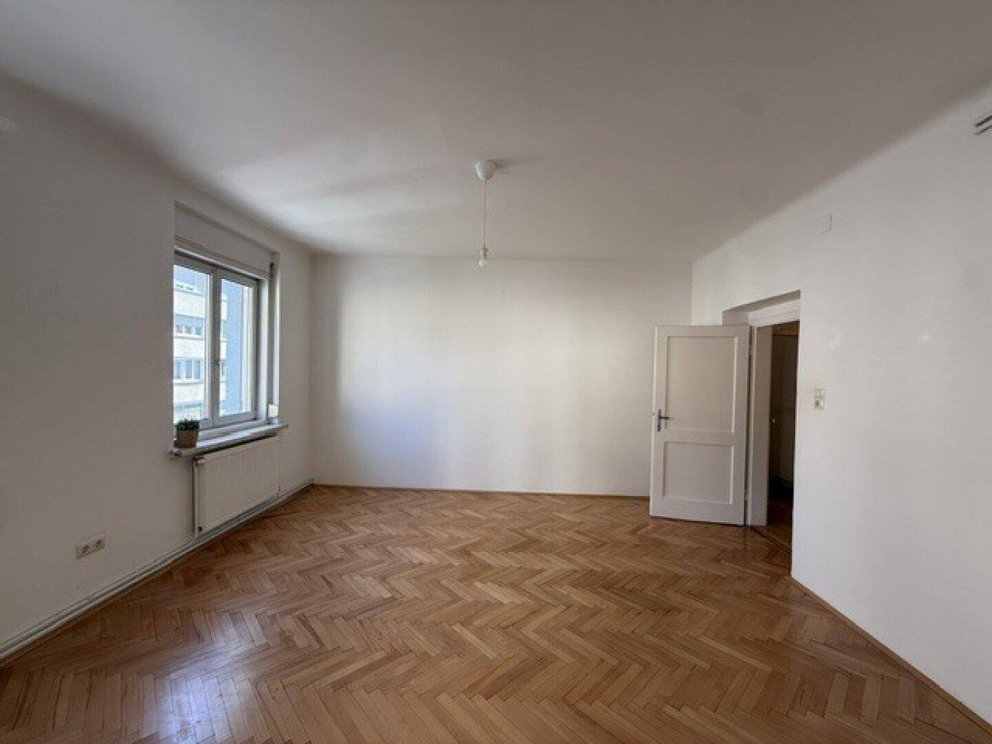 Heller, leerer Raum mit klassischem Fischgrätparkett und einem Fenster mit Blick nach draußen.