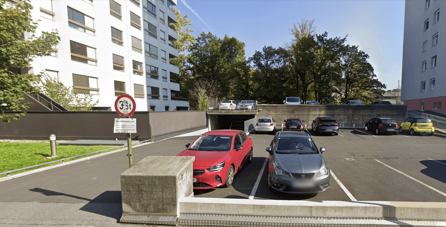 Außenansicht des Gebäudes mit Tiefgaragenzufahrt und oberirdischen Parkplätzen unter sonnigem Himmel.