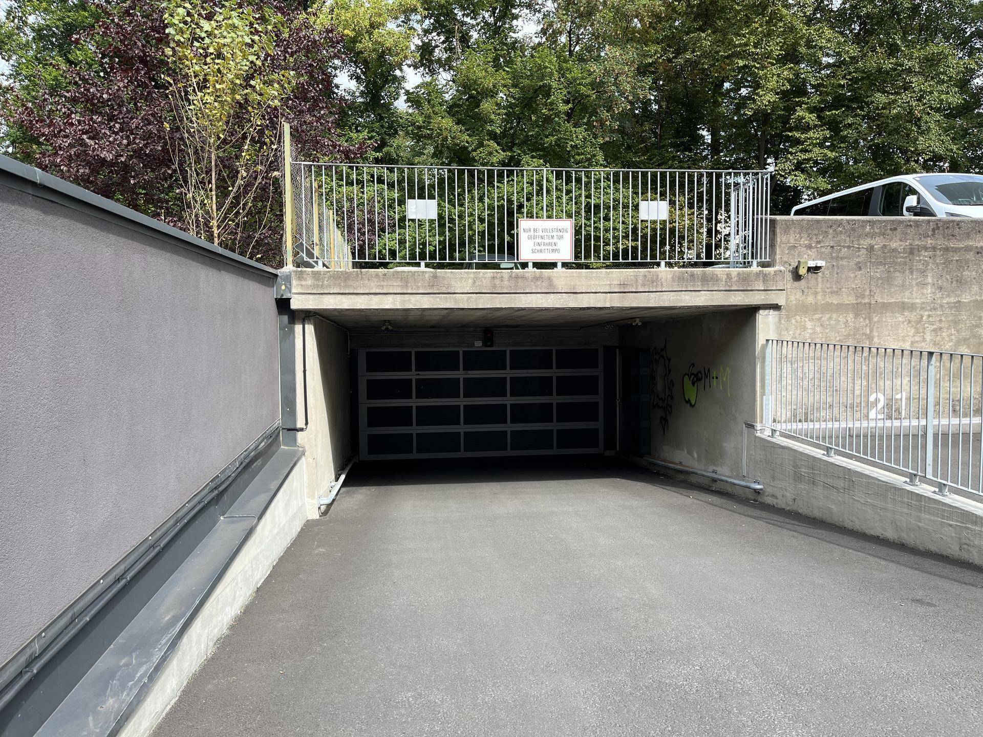 Zufahrt zur Tiefgarage mit Betonwänden, Metallgeländer und umgebender Vegetation unter blauem Himmel.
