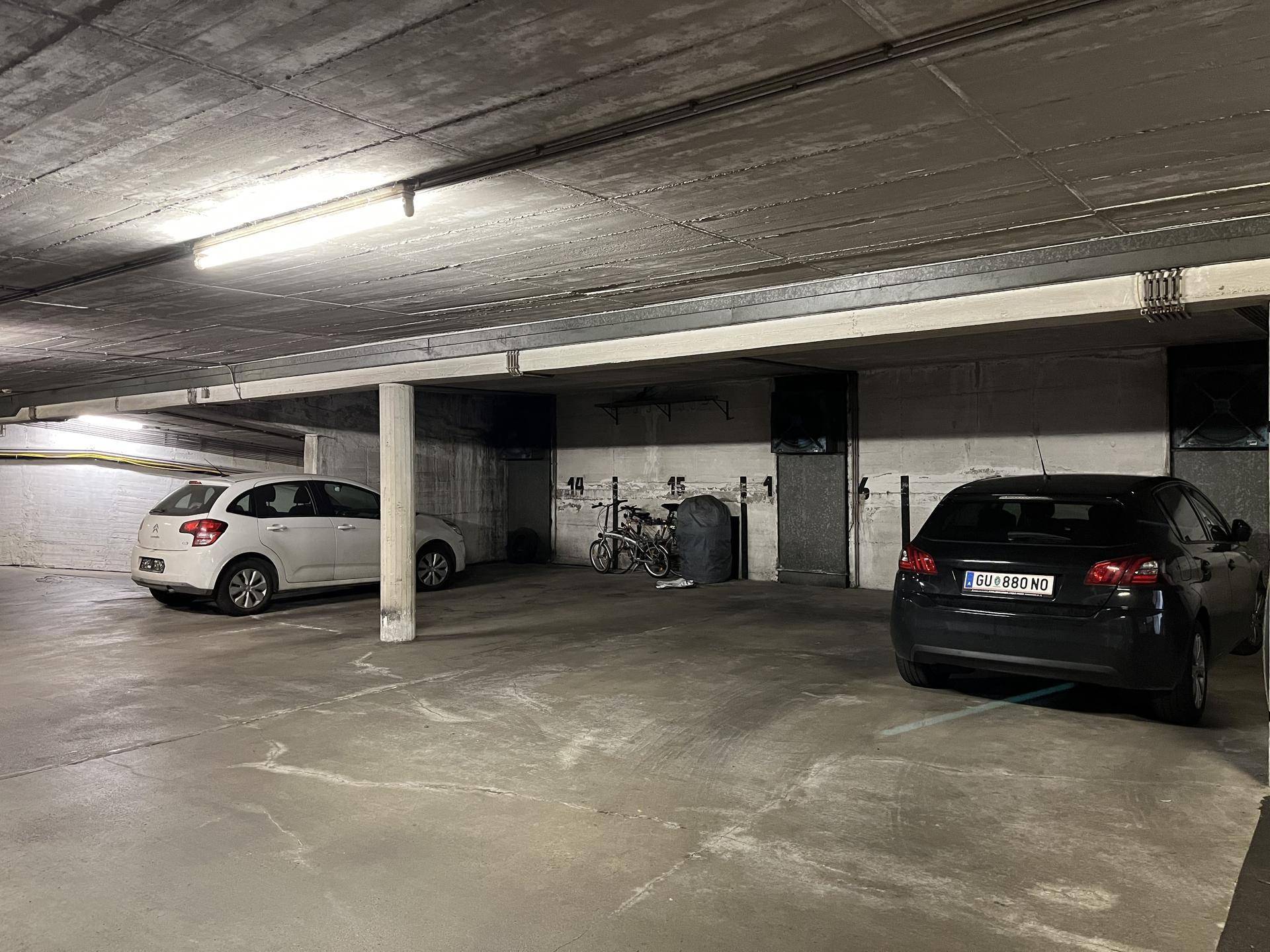 Geräumige Tiefgarage mit zwei geparkten Autos, Fahrrädern und sichtbaren Leuchtstoffröhren an der Decke.