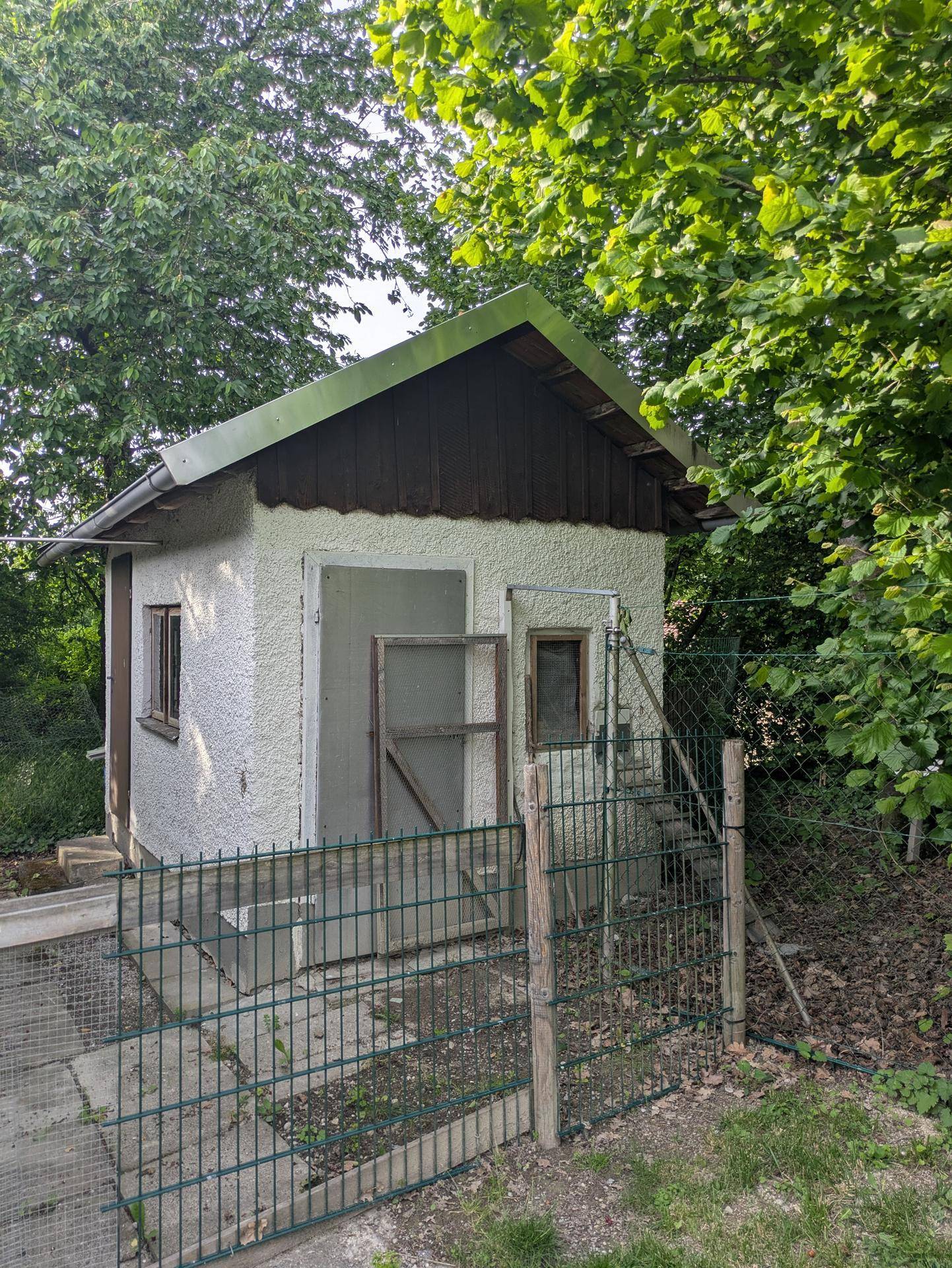 Kleines Gartenhaus mit einfacher Bauweise, umgeben von üppigem Grün und Bäumen.