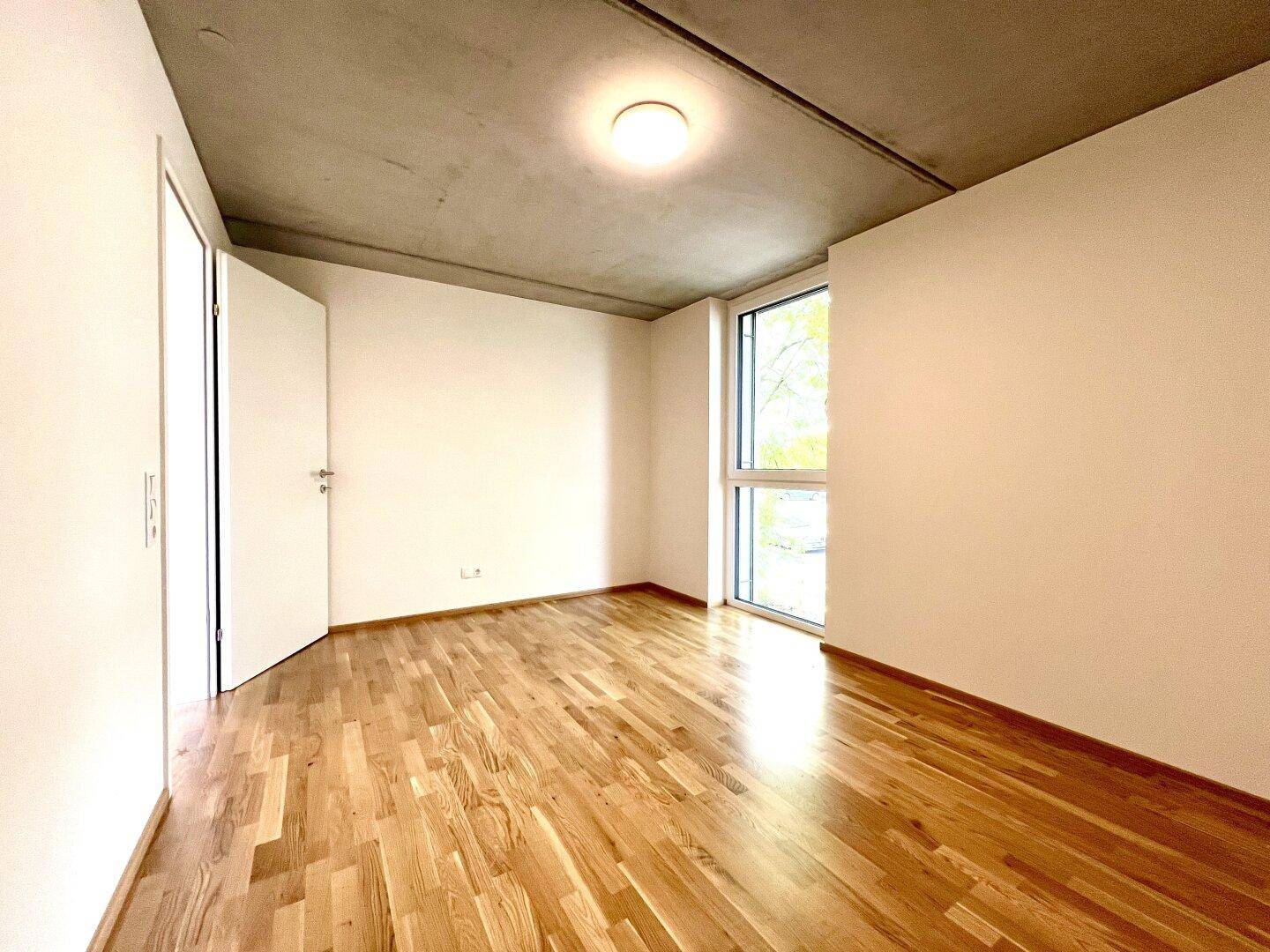 Helles Schlafzimmer mit Parkettboden und einem großen Fenster, das viel Tageslicht hereinlässt.