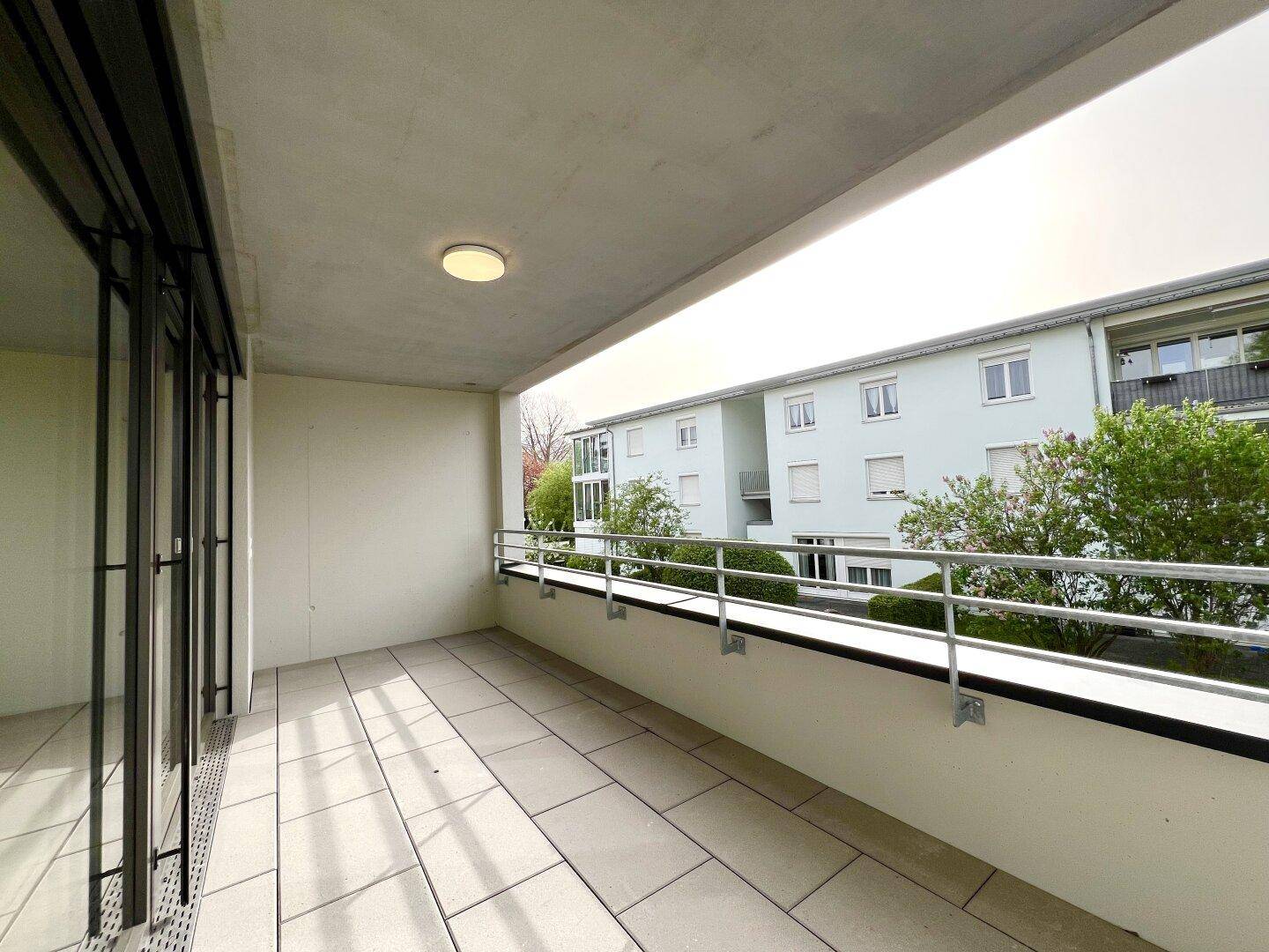 Heller Balkon mit Fliesenboden und Metallgeländer, bietet einen Blick auf die umliegenden Gebäude und Bäume.