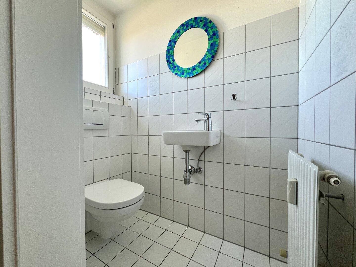 Separates WC mit Toilette, kleinem Waschbecken, Fenster und dekorativem Spiegel.